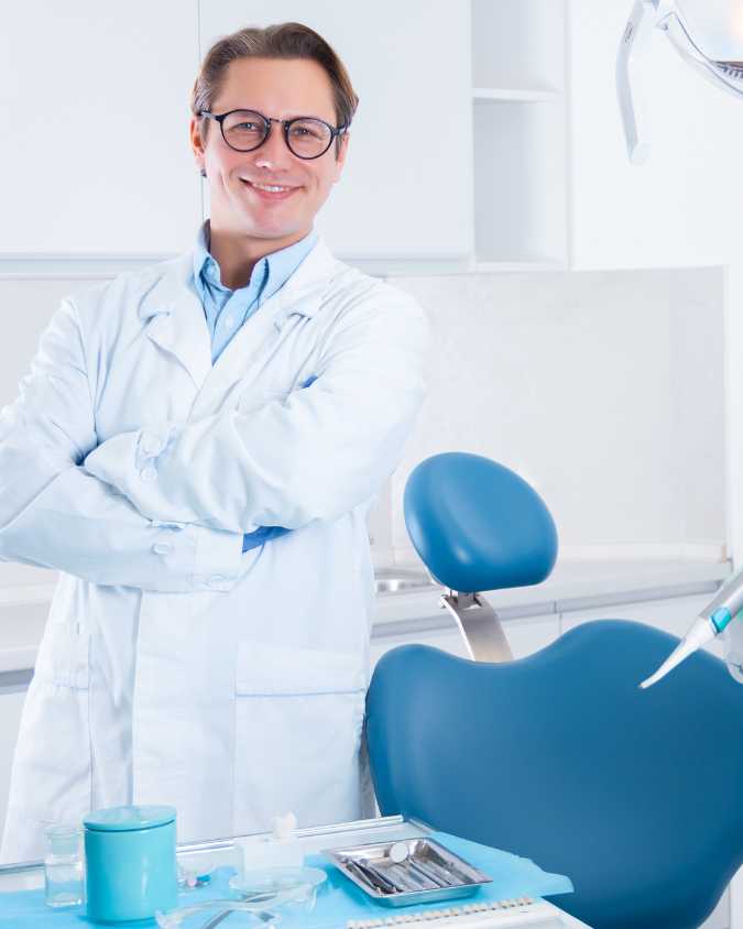 White male in white lab coat wearing glasses with brown hair, next to dental chair with dentist instruments.
