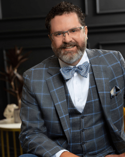White male, with graying beard, and dark curly hair, wear glasses, jeans, a suit jacket and vest, with a white shirt and blue bow tie