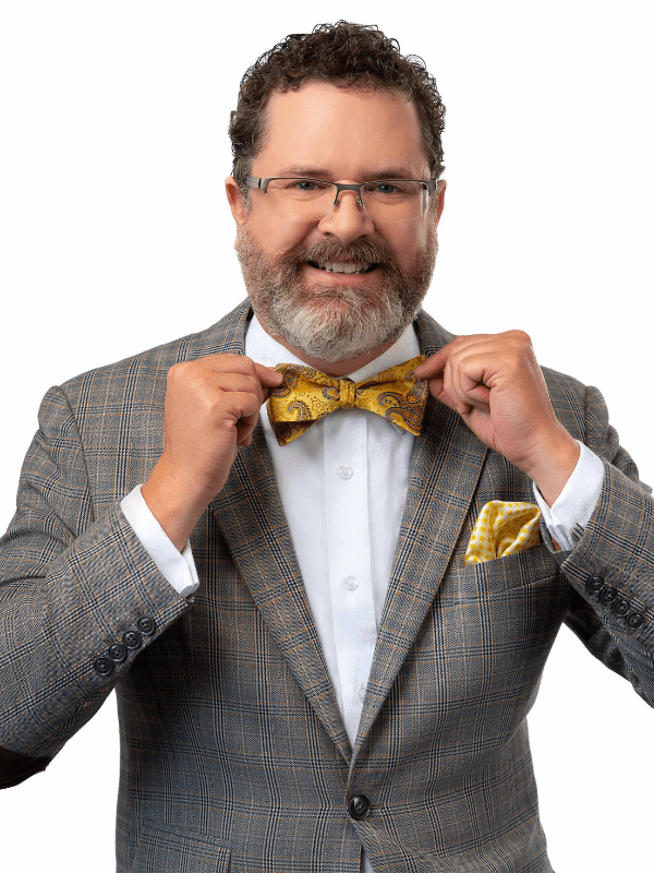 White male, with graying beard, and dark curly hair, wear glasses, a gray suit jacket , with a white shirt and yellow bow tie